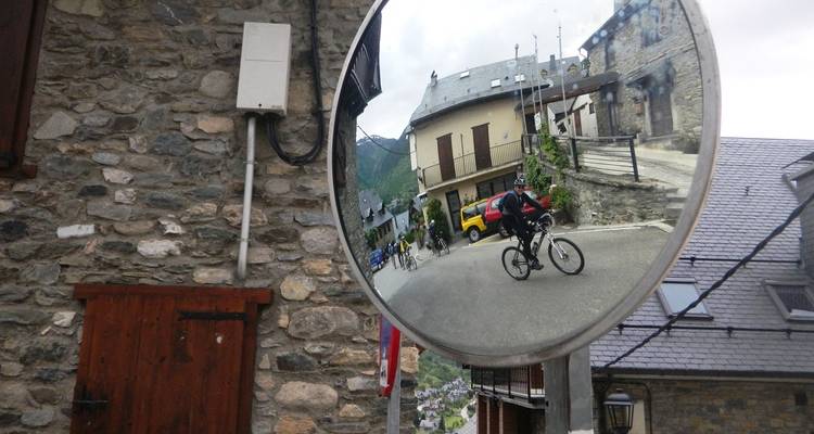 On voit un cycliste dans le reflet d'un miroir de rue convexe installé sur un bâtiment en pierre dans un village de montagne.