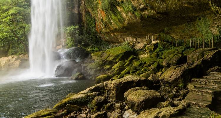 Poderosa cascada se precipita en una piscina neblinosa junto a rocas cubiertas de musgo bajo un acantilado saliente