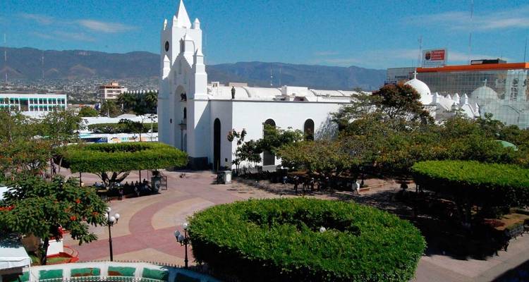 Iglesia blanca de estilo colonial dominando una plaza frondosa con montañas de fondo