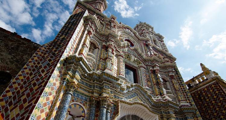 Perspectiva hacia arriba de una fachada ornamentada de iglesia barroca mexicana ricamente decorada con azulejos coloridos contra un cielo azul.