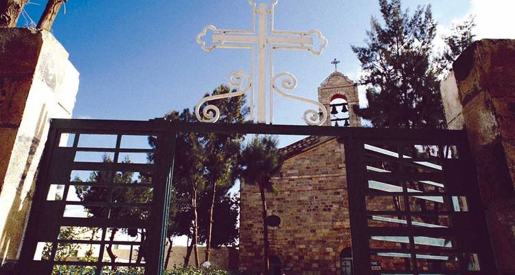 Metal gate with ornate cross frames a stone church and bell tower under a bright blue sky.