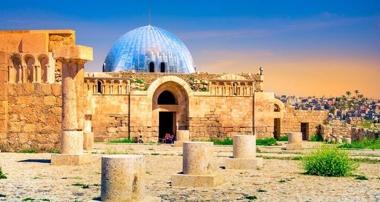 Stone courtyard and domed Umayyad Palace building within ancient citadel ruins under a colorful sunset sky.