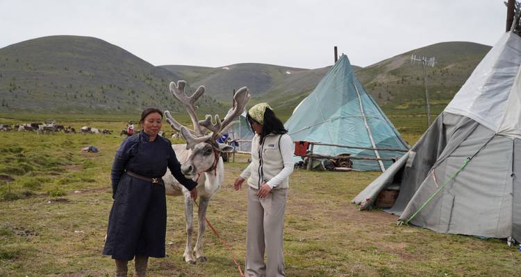 Dos mujeres de pie con un reno frente a tiendas de campaña en un paisaje montañoso.