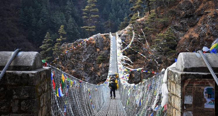 Gente cruzando un puente colgante decorado con banderas de oración.