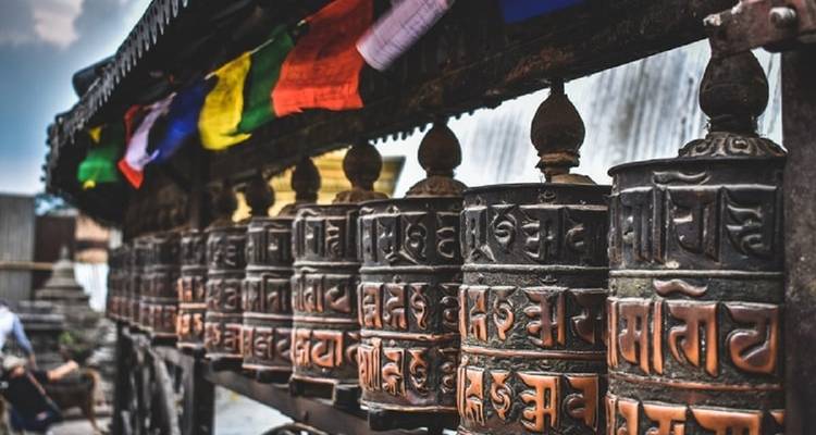 Row of prayer wheels with colorful flags in the background.
