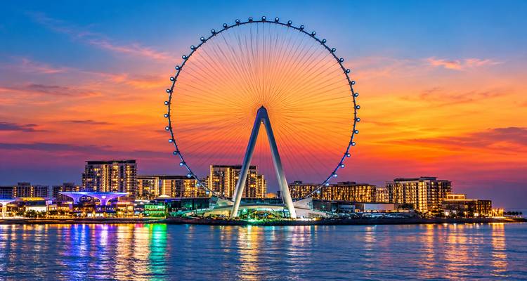 La gigantesca rueda de observación Ain Dubai iluminada contra un cielo al atardecer vibrante reflejada en las tranquilas aguas del golfo.