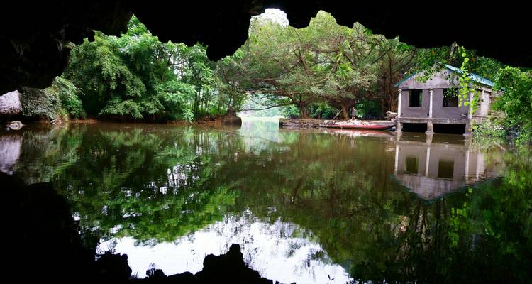 Blick aus dem Inneren einer Flusshöhle hinaus auf ruhiges Wasser, das üppige Bäume und eine kleine Hütte am Wasser widerspiegelt