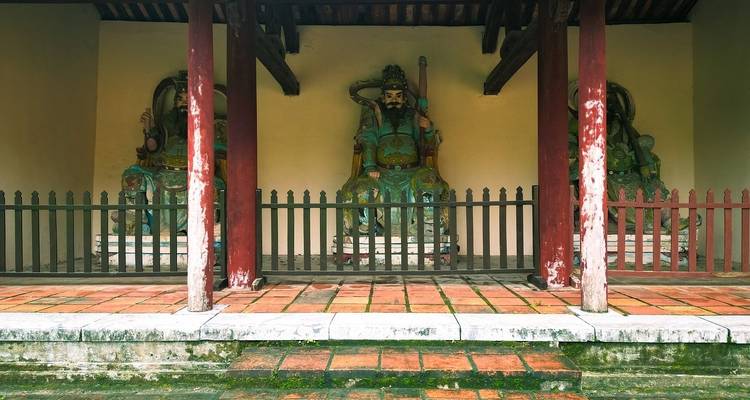 Drei verwitterte Wächterstatuen stehen hinter hölzernen Geländern in einer historischen vietnamesischen Tempelhalle.