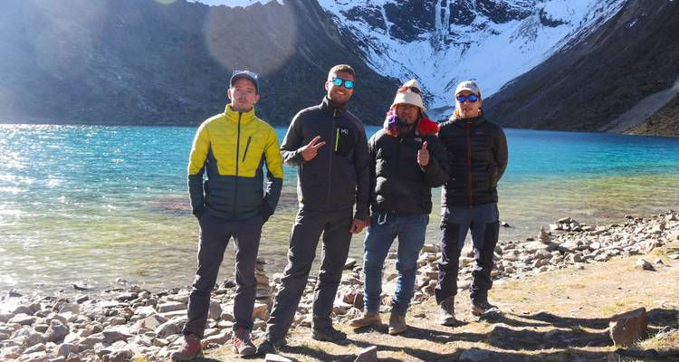 Vier Personen posieren an einem türkisfarbenen See mit Bergen im Hintergrund.