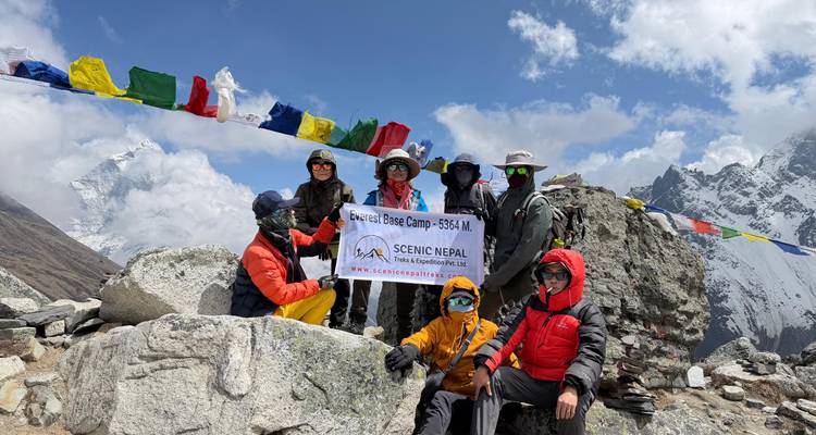 Trekking-Team posiert mit einem Banner des Everest Base Camp zwischen Gebetsfahnen und verschneiten Himalaya-Gipfeln.
