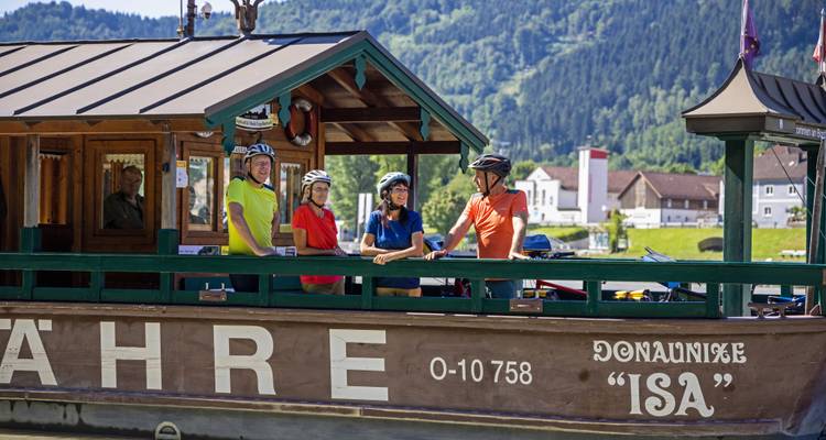 Des cyclistes portant des casques debout sur un bateau-ferry en bois nommé « ISA » traversant un large fleuve européen.