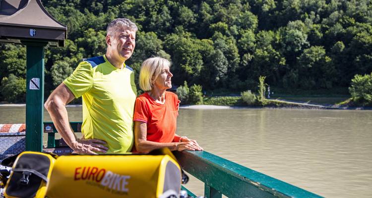 Couple de personnes d'âge mûr vêtues de tenues de cyclisme colorées s'appuyant sur la rambarde d'un traversier, admirant le paysage fluvial serein.