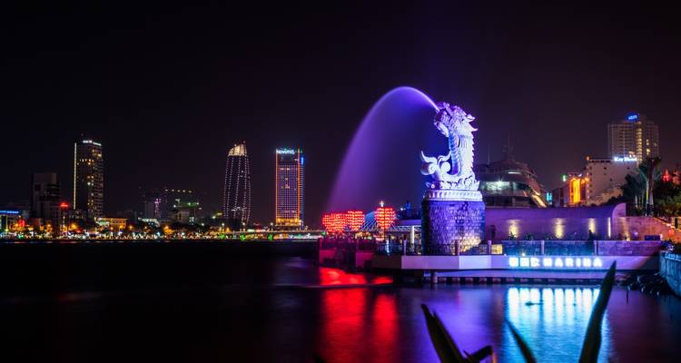 Night view of Da Nang waterfront with the illuminated carp-dragon statue spraying water and city skyline in neon lights.