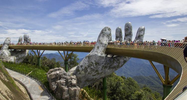 The Golden Bridge in Ba Na Hills supported by giant stone hands, crowded with visitors above mountain scenery.