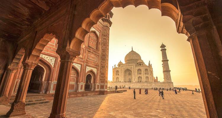 Vista de la hora dorada del Taj Mahal enmarcado por el arco ornamentado de la mezquita con visitantes en el patio.