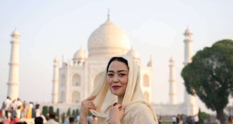 Mujer elegante con pañuelo en la cabeza sonriendo frente al Taj Mahal suavemente iluminado al anochecer.