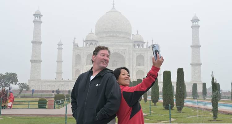 Couple takes a selfie with the misty Taj Mahal rising in the background on a foggy morning.