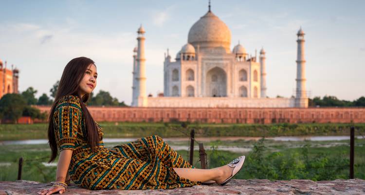 Young woman in patterned dress poses on a stone ledge with sunset light on the Taj Mahal behind her.
