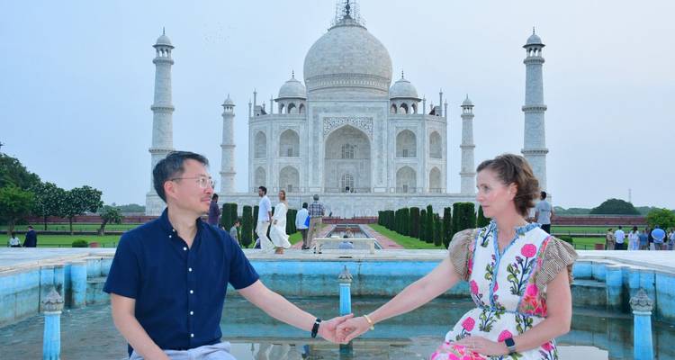 Couple sit hand-in-hand on the marble pool edge leading to the Taj Mahal at dusk.