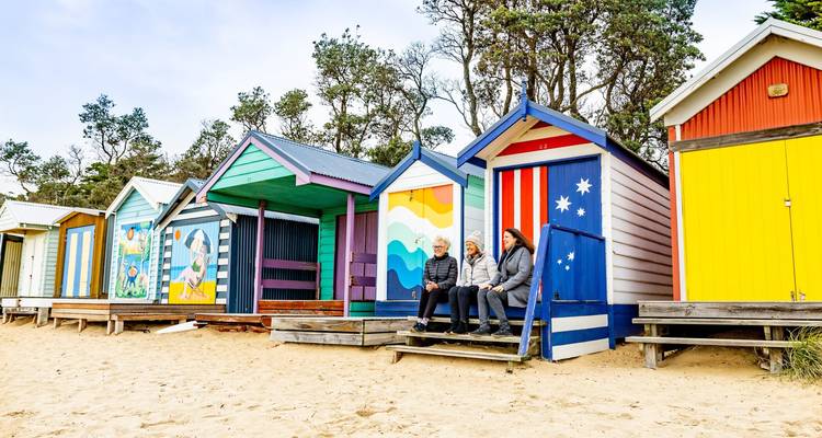 Fila de casetas de playa pintadas de colores brillantes con tres amigos charlando en los escalones.