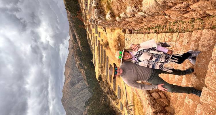 Pareja usando sombreros y ponchos de pie en terrazas incas con dramáticas montañas andinas detrás.