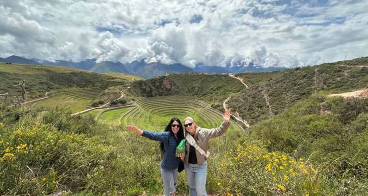 Two smiling women posing with arms raised before the circular agricultural terraces of Moray in the Andes