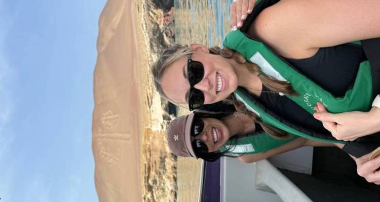 Two women in life vests smiling on a speedboat with the Paracas Candelabra geoglyph visible on the desert hillside behind