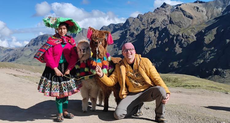 Une femme vêtue de couleurs vives et des alpagas avec des montagnes en arrière-plan.