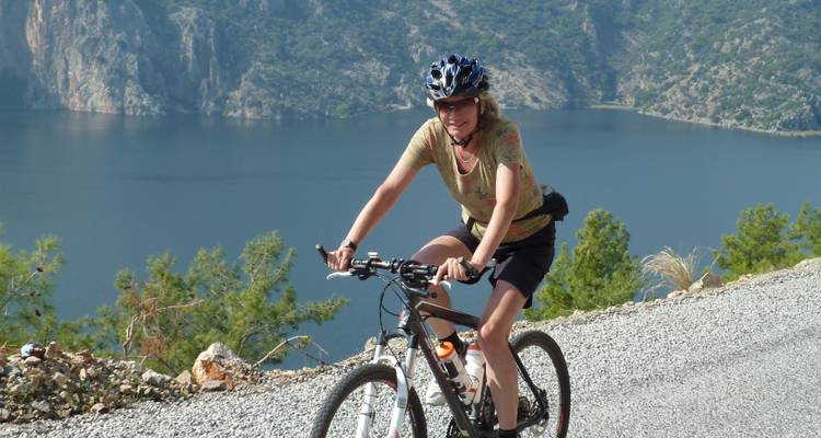 A cyclist riding along a lakeside road with mountains.