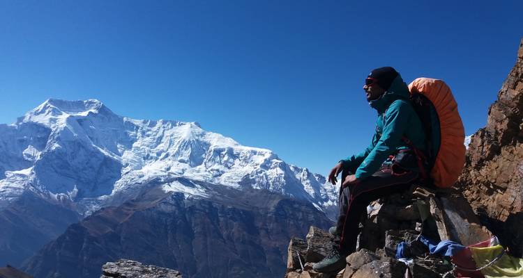 Person, die auf einem Bergsims sitzt, mit schneebedeckten Gipfeln im Hintergrund.