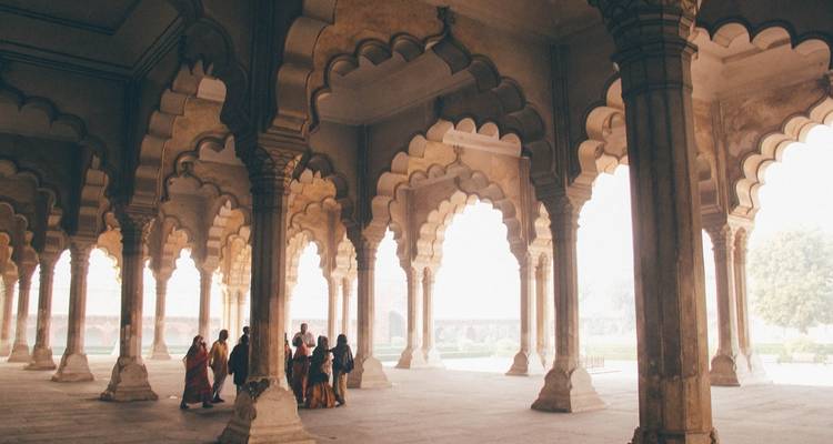 Visitors in colorful saris gather under the scalloped arches of a sandstone hall filled with soft, diffused morning light.