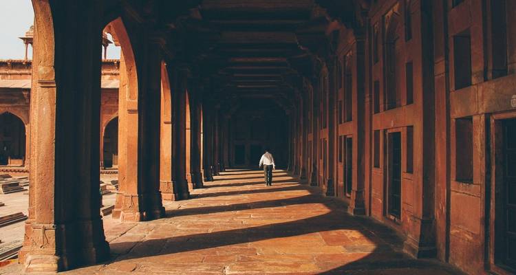 A lone visitor walks through a shadowed sandstone corridor lined with columns inside a Mughal fort.