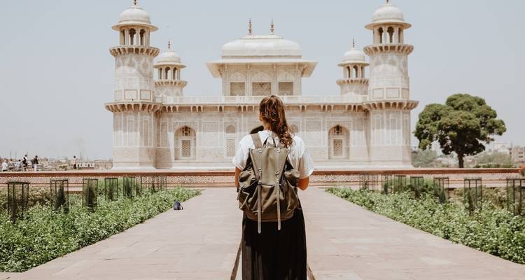 A backpacked traveler stands facing the ornate Baby Taj, framed by lush green gardens on a sunny day.