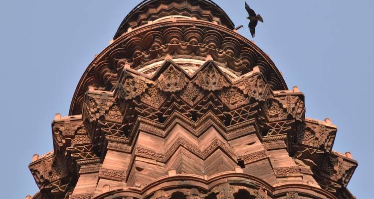 Close-up of the intricately carved crown of Qutub Minar with a bird soaring through the clear sky.