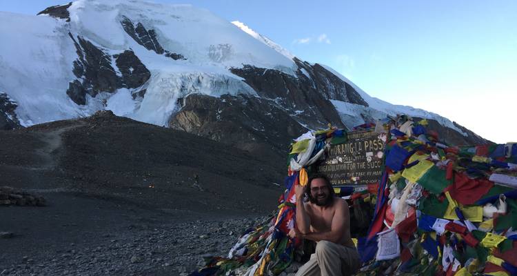 Smiling man at Thorong La Pass with prayer flags and mountains in the background.