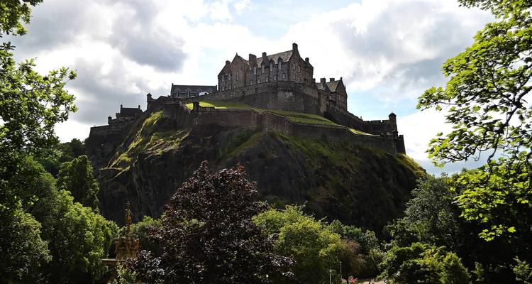 Edinburgh Castle on a hill surrounded by trees and cloudy sky.