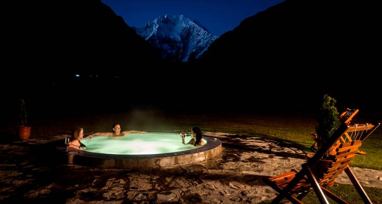 Drei Reisende entspannen sich nachts in einem Außen-Whirlpool, während ein schneebedeckter Gipfel dahinter in Silhouette erkennbar ist.