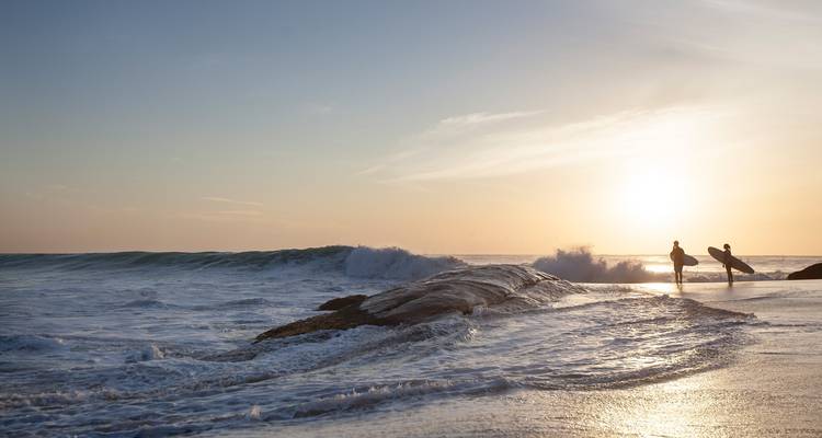 Twee surfers die langs het water lopen met een zonsondergang op de achtergrond.
