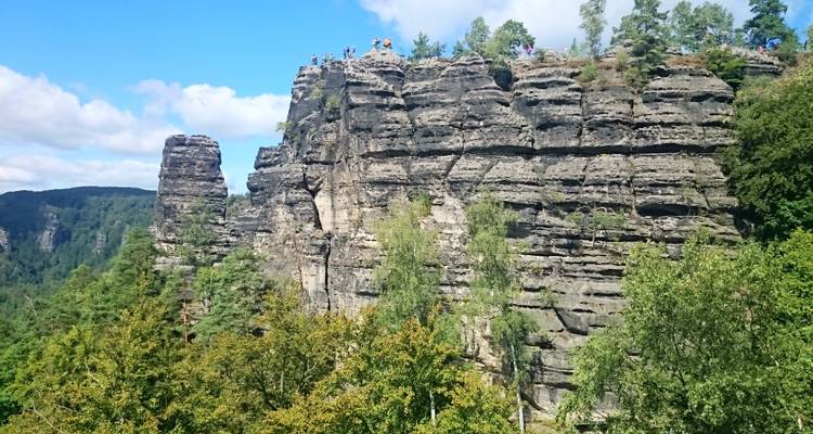 Rock cliff with people standing on top, surrounded by forest.