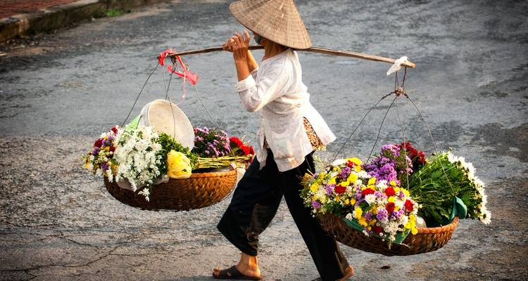Vendor carrying baskets of flowers on a bustling street.
