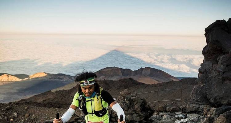Trail runner climbing a volcanic terrain with clouds over mountains.