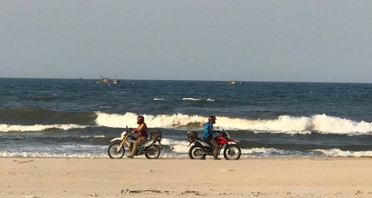 Two motorcyclists riding on a beach with ocean waves.