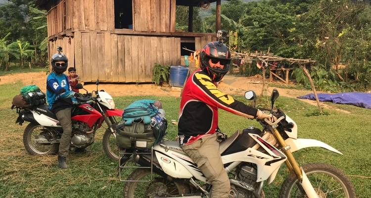 Two motorcyclists near a rustic house with green surroundings.