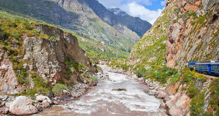Train travels alongside a river with rocky cliffs.
