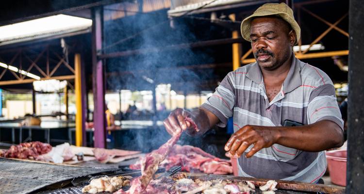 A man grilling meat at a market stall with smoke rising.