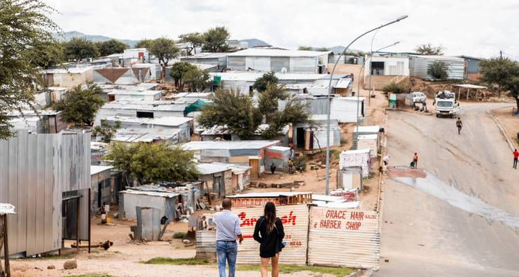Two people walking through an informal settlement with corrugated metal structures.