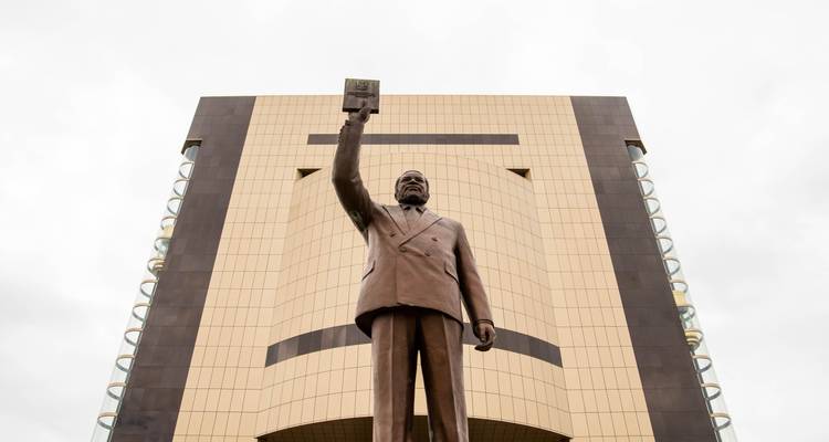 A statue in front of a modern building, the statue holds an item in the air.