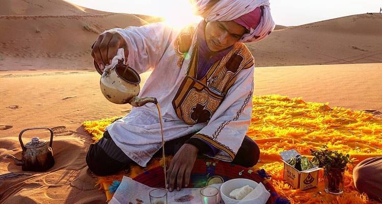 Person pouring traditional tea on a vibrant orange blanket in the desert.