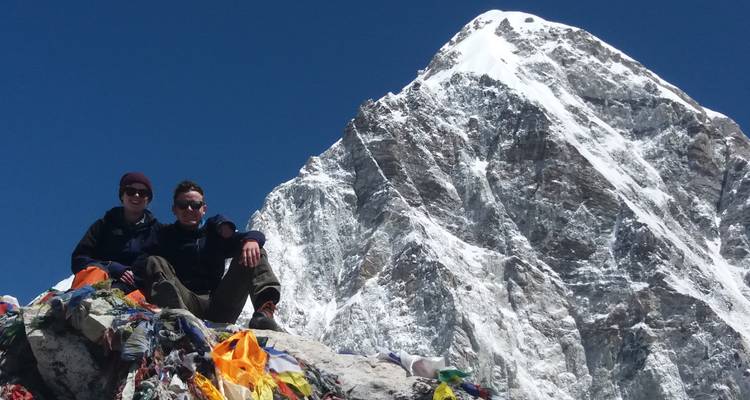 Two people sitting among colorful flags in front of a snowy peak.
