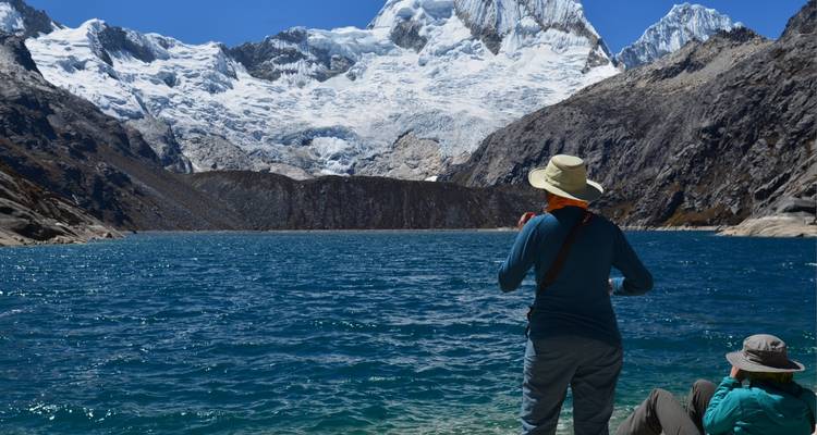 Person standing by a turquoise alpine lake with snow-covered mountains in the background.